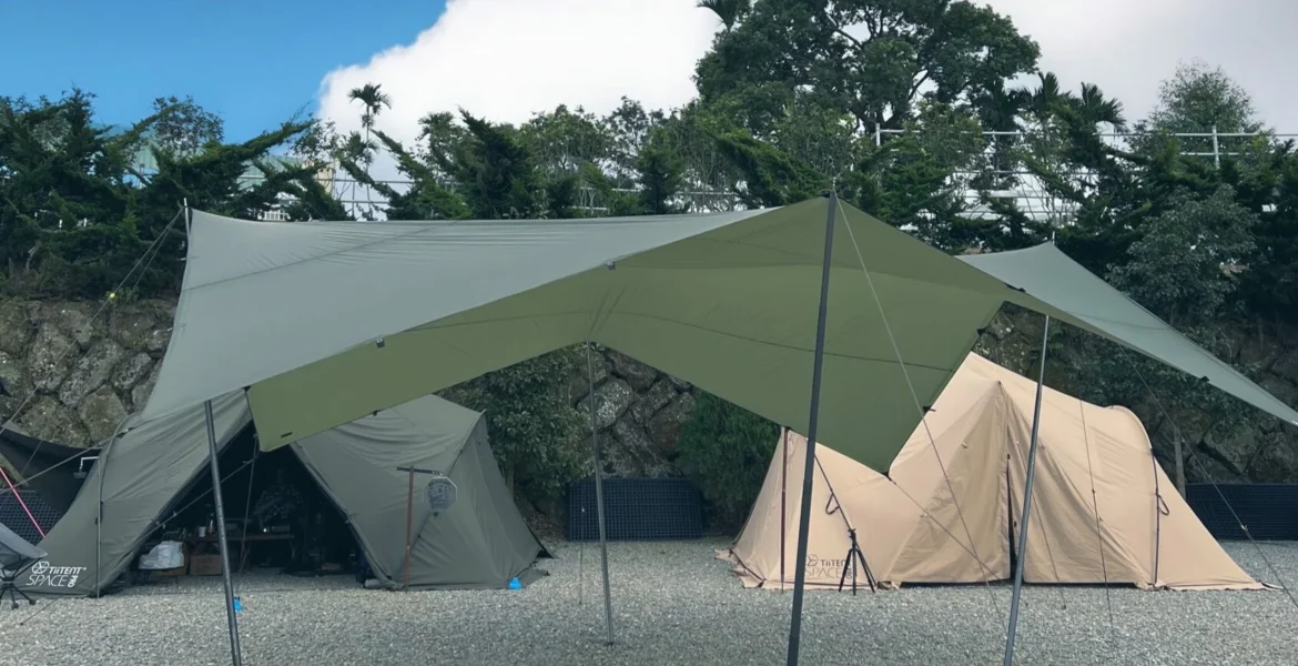 在碎石營地上搭建的軍綠色大型六角天幕，背景可見山區植被與淡黃色隧道帳篷，展示台灣露營常見的遮陽天幕佈局。