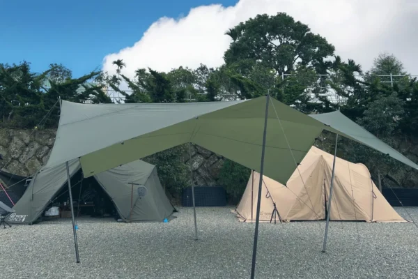 在碎石營地上搭建的軍綠色大型六角天幕，背景可見山區植被與淡黃色隧道帳篷，展示台灣露營常見的遮陽天幕佈局。